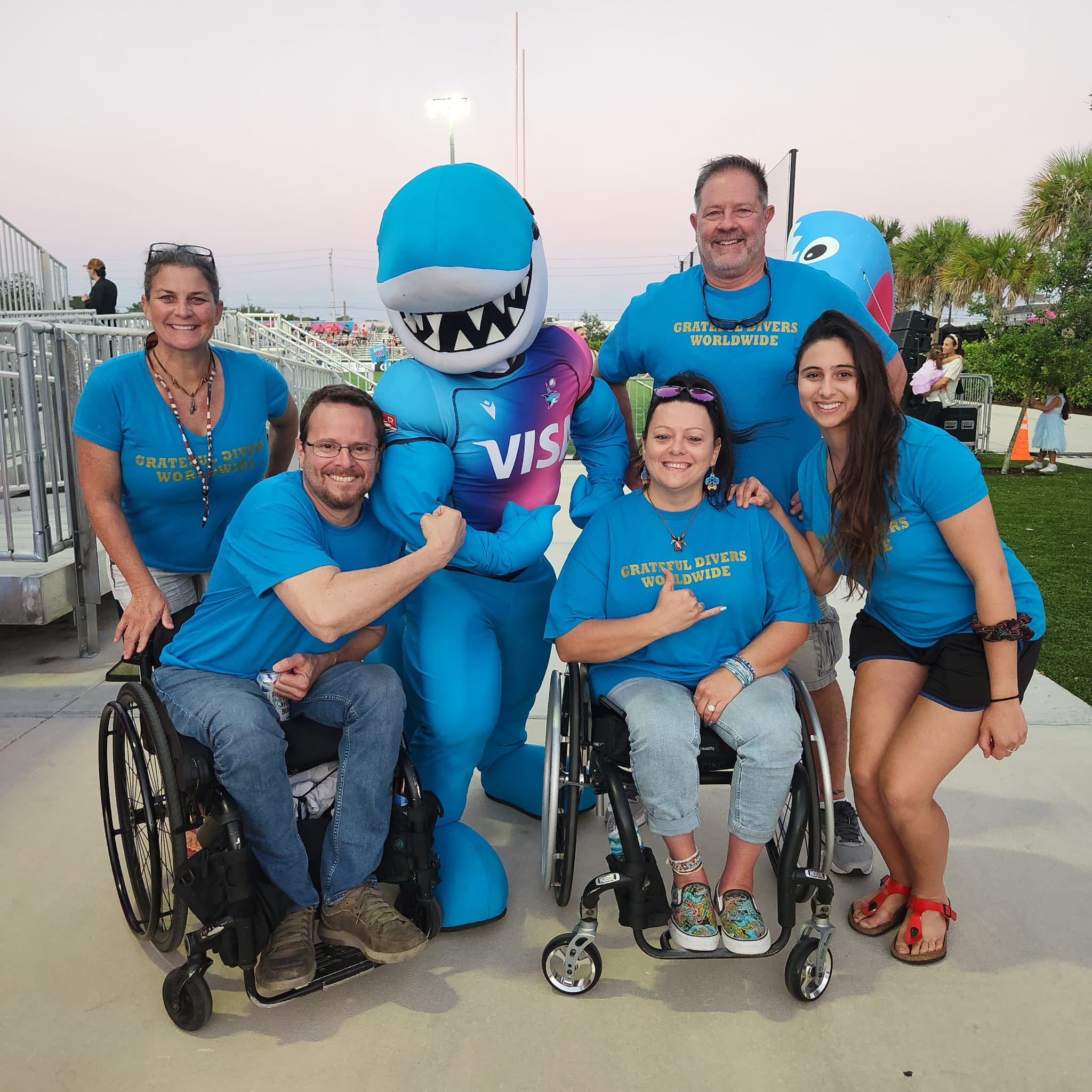 A group of six smiling individuals wearing matching blue “Grateful Divers Worldwide” shirts pose with a friendly shark mascot in a brightly colored VISA wetsuit. Two members of the group are in wheelchairs. The photo is taken outdoors during sunset at an event, with stadium seating and palm trees in the background.