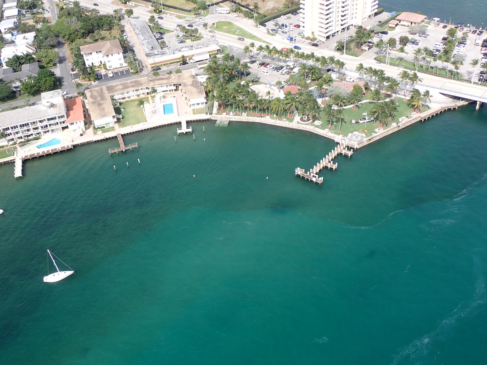Aerial view of the shoreline near Wahoo Bay in Pompano Beach, Florida. The image shows waterfront condos with pools, a green park lined with palm trees, boat docks extending into the clear turquoise water, and a sailboat anchored nearby. A bridge and parking lot are visible at the top right.