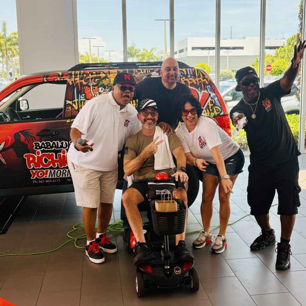 Scooter Bobby seated on a mobility scooter, surrounded by friends and radio station crew in front of a colorful Yo! 107.1 promotional van, all smiling and striking playful poses.
