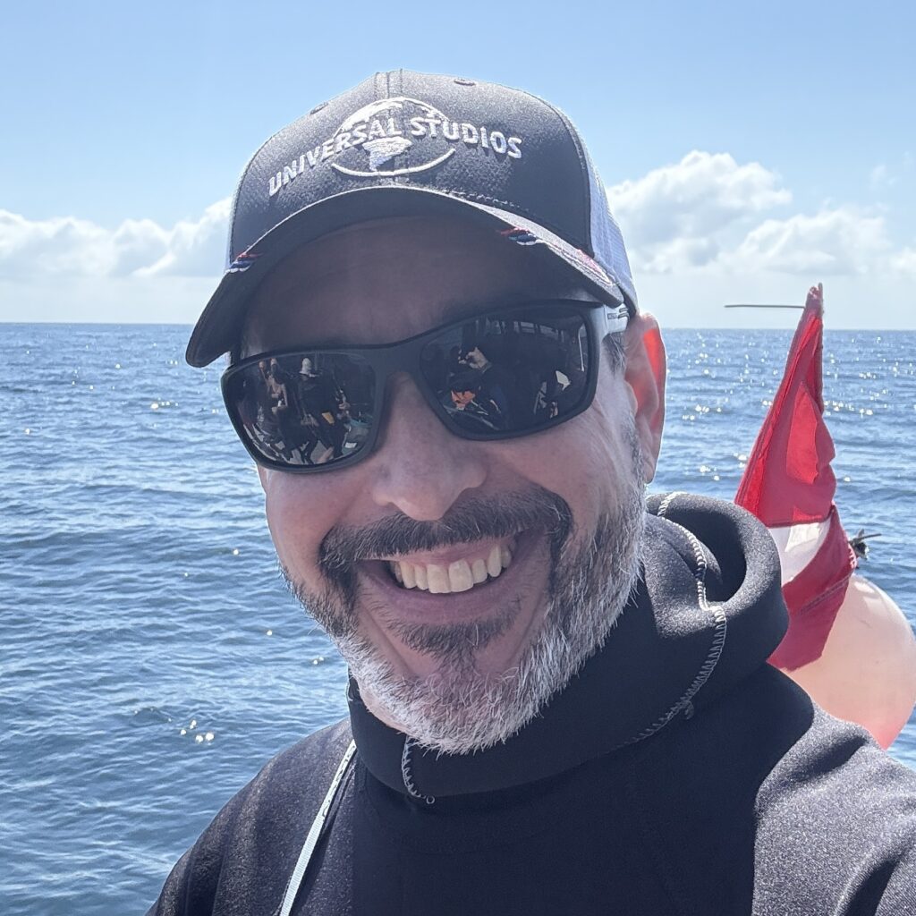 Scooter Bobby smiling on a dive boat in the sunshine, wearing a Universal Studios cap and sunglasses, with the ocean and a dive flag behind him.
