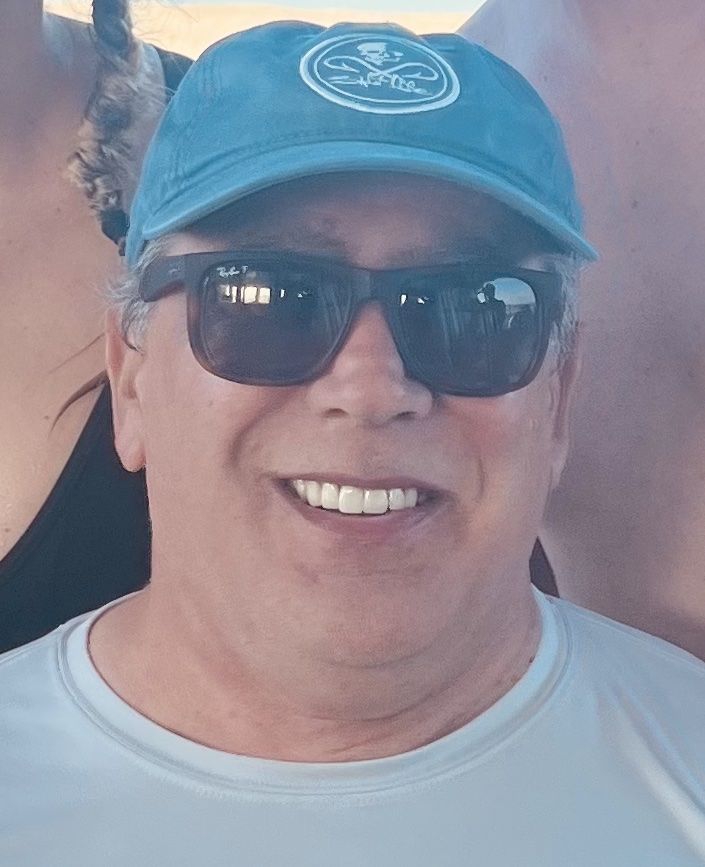 Close-up of Todd smiling in a light blue shirt, sunglasses, and a teal baseball cap with a white logo, taken in a sunny outdoor setting.